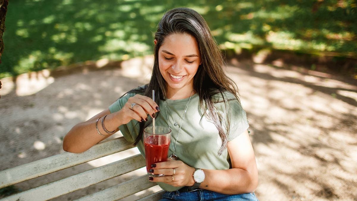 A happy young woman relaxes on a park bench with refreshing juice, enjoying a sunny day outdoors.